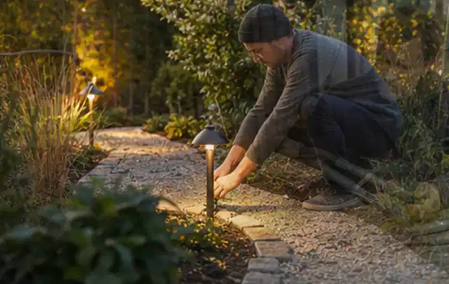 Person richtet eine niedrige warm leuchtende Gartenlampe am Kiesweg aus; Thema Mähroboter für kleine Gärten, Schwerpunkt Ersatzteile; Praxisalltag.