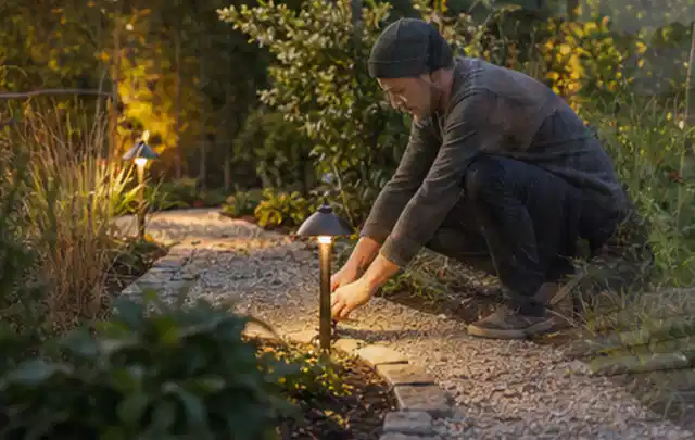 Person richtet eine niedrige warm leuchtende Gartenlampe am Kiesweg aus; Thema Mähroboter für kleine Gärten, Schwerpunkt Fehlerhilfe; Ausgangslage.