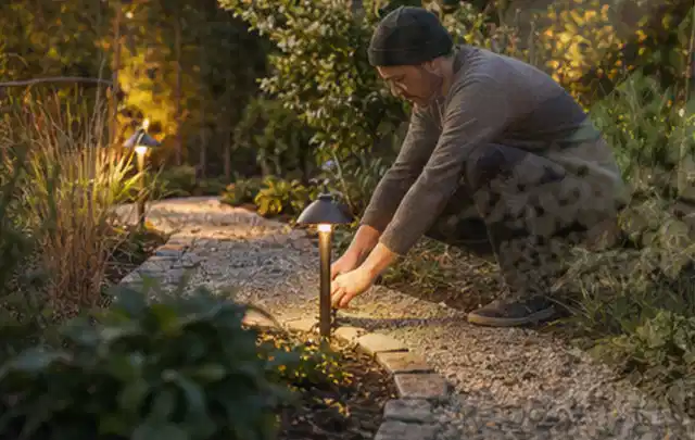Person richtet eine niedrige warm leuchtende Gartenlampe am Kiesweg aus; Thema Mähroboter für kleine Gärten, Schwerpunkt Leistung; Ausgangslage.