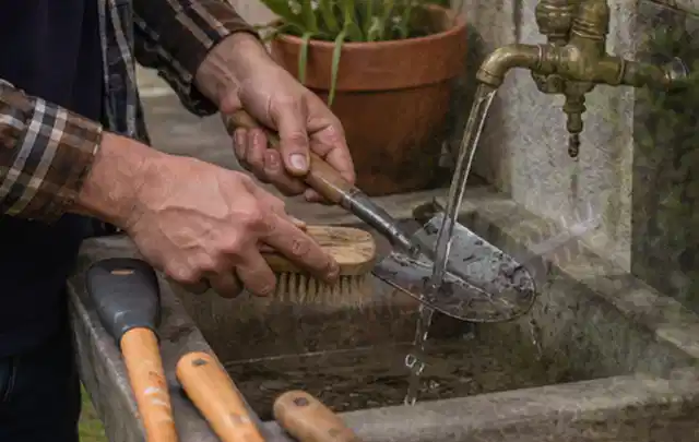 Hände reinigen gebrauchte Gartengeräte mit Bürste und Wasser am Außenbecken; Thema Mähroboter für kleine Gärten, Schwerpunkt Leistung; Praxisalltag.