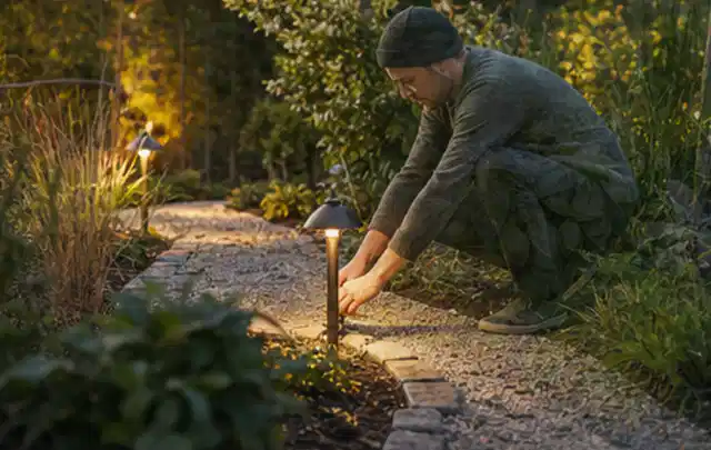 Person richtet eine niedrige warm leuchtende Gartenlampe am Kiesweg aus; Thema Mähroboter für kleine Gärten, Schwerpunkt Reinigung; Praxisalltag.
