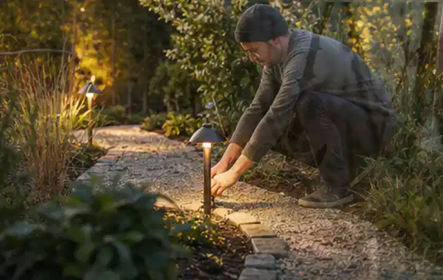 Person richtet eine niedrige warm leuchtende Gartenlampe am Kiesweg aus; Thema Mähroboter für kleine Gärten, Schwerpunkt Systemvergleich; Ausgangslage.
