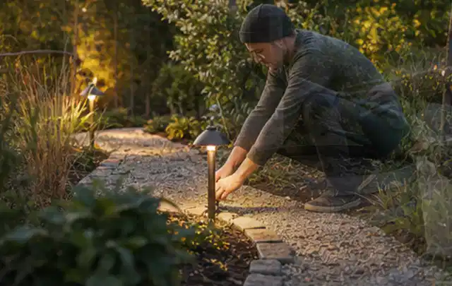Person richtet eine niedrige warm leuchtende Gartenlampe am Kiesweg aus; Thema Mähroboter für kleine Gärten, Schwerpunkt Testkriterien; Ausgangslage.