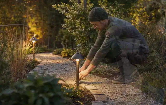 Person richtet eine niedrige warm leuchtende Gartenlampe am Kiesweg aus; Thema Mähroboter für kleine Gärten, Schwerpunkt Vergleich; Praxisalltag.