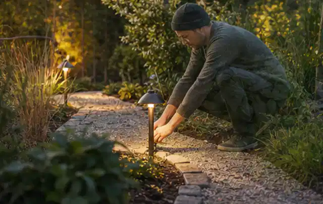Person richtet eine niedrige warm leuchtende Gartenlampe am Kiesweg aus; Thema Mähroboter für kleine Gärten, Schwerpunkt Wartung; Praxisalltag.