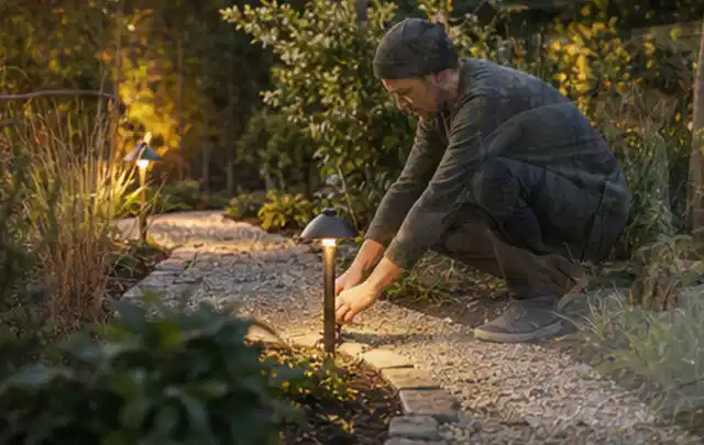 Person richtet eine niedrige warm leuchtende Gartenlampe am Kiesweg aus; Thema Mähroboter Garagen, Schwerpunkt Reinigung; Kontrolle.
