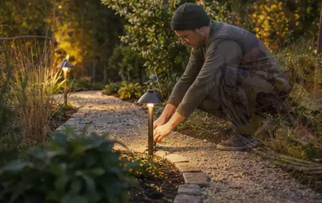 Person richtet eine niedrige warm leuchtende Gartenlampe am Kiesweg aus; Thema Mähroboter Garagen, Schwerpunkt Vergleich; Kontrolle.