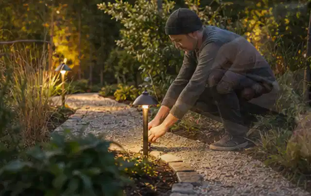 Person richtet eine niedrige warm leuchtende Gartenlampe am Kiesweg aus; Thema Mähroboter Garagen, Schwerpunkt Wartung; Umsetzung.