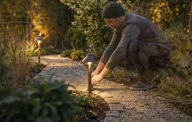 Person richtet eine niedrige warm leuchtende Gartenlampe am Kiesweg aus; Thema Mähroboter Garagen, Schwerpunkt Winterlager; Ausgangslage.