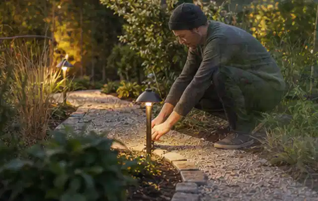 Person richtet eine niedrige warm leuchtende Gartenlampe am Kiesweg aus; Thema Mähroboter Installation, Schwerpunkt Systemvergleich; Ergebnis.