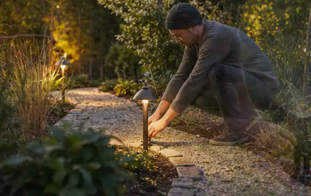 Person richtet eine niedrige warm leuchtende Gartenlampe am Kiesweg aus; Thema Mähroboter Messer, Schwerpunkt Anwendung; Praxisalltag.