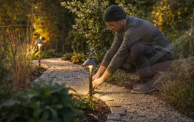 Person richtet eine niedrige warm leuchtende Gartenlampe am Kiesweg aus; Thema Mähroboter Messer, Schwerpunkt Kaufberatung; Ergebnis.
