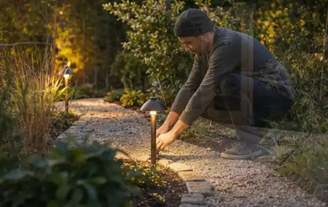 Person richtet eine niedrige warm leuchtende Gartenlampe am Kiesweg aus; Thema Mähroboter Messer, Schwerpunkt Mähroboter; Umsetzung.