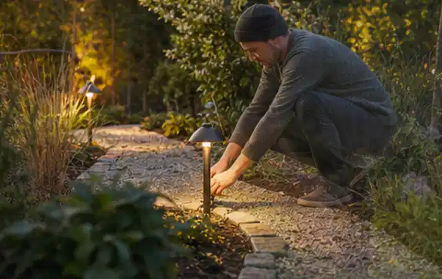 Person richtet eine niedrige warm leuchtende Gartenlampe am Kiesweg aus; Thema Mähroboter Messer, Schwerpunkt Nachhaltigkeit; Praxisalltag.