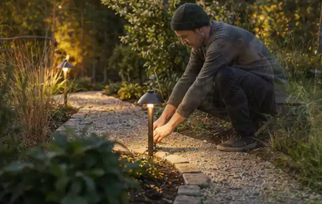Person richtet eine niedrige warm leuchtende Gartenlampe am Kiesweg aus; Thema Mähroboter Messer, Schwerpunkt Sicherheit; Umsetzung.