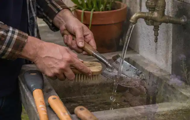 Hände reinigen gebrauchte Gartengeräte mit Bürste und Wasser am Außenbecken; Thema Mähroboter Messer, Schwerpunkt Testkriterien; Umsetzung.