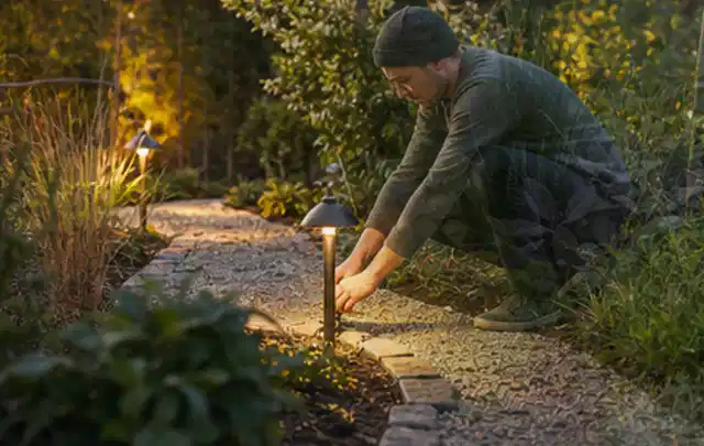 Person richtet eine niedrige warm leuchtende Gartenlampe am Kiesweg aus; Thema Mähroboter Messer, Schwerpunkt Vergleich; Umsetzung.