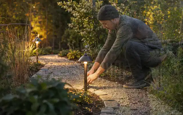 Person richtet eine niedrige warm leuchtende Gartenlampe am Kiesweg aus; Thema Mähroboter Messer, Schwerpunkt Wartung; Praxisalltag.