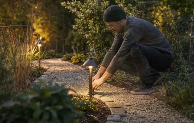 Person richtet eine niedrige warm leuchtende Gartenlampe am Kiesweg aus; Thema Mähroboter mit GPS, Schwerpunkt Dimensionierung; Ausgangslage.