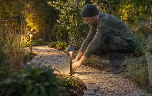 Person richtet eine niedrige warm leuchtende Gartenlampe am Kiesweg aus; Thema Mähroboter mit GPS, Schwerpunkt Leistung; Ergebnis.