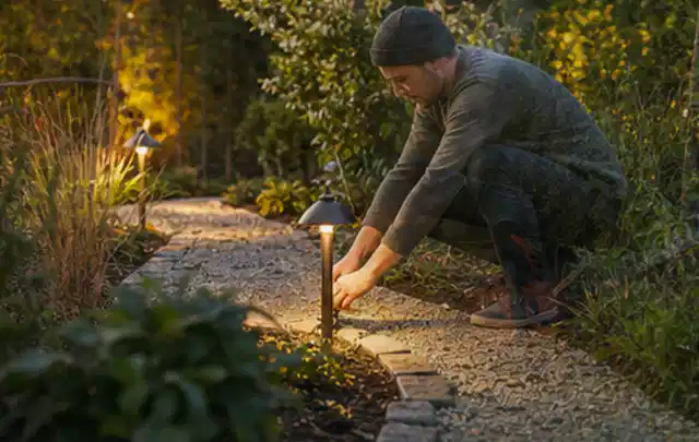 Person richtet eine niedrige warm leuchtende Gartenlampe am Kiesweg aus; Thema Mähroboter mit GPS, Schwerpunkt Mähroboter; Umsetzung.