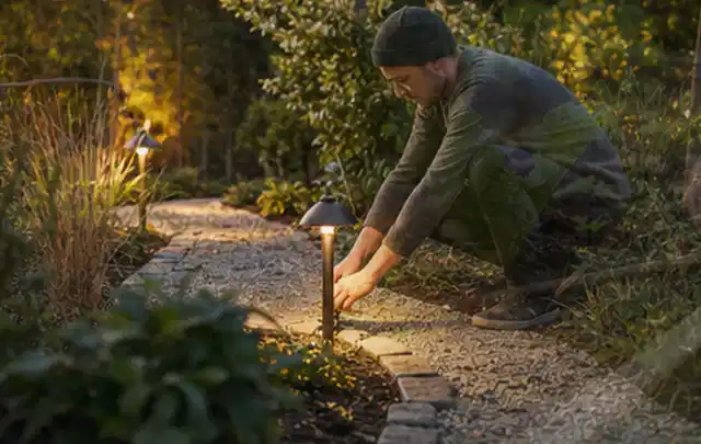 Person richtet eine niedrige warm leuchtende Gartenlampe am Kiesweg aus; Thema Mähroboter mit GPS, Schwerpunkt Nachhaltigkeit; Praxisalltag.