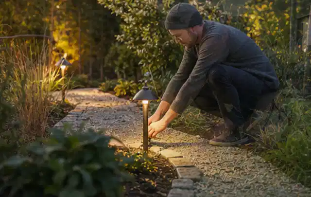 Person richtet eine niedrige warm leuchtende Gartenlampe am Kiesweg aus; Thema Mähroboter mit GPS, Schwerpunkt Winterlager; Umsetzung.