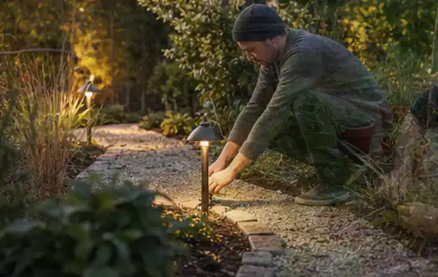 Person richtet eine niedrige warm leuchtende Gartenlampe am Kiesweg aus; Thema Mähroboter ohne Begrenzungskabel, Schwerpunkt Anwendung; Ausgangslage.