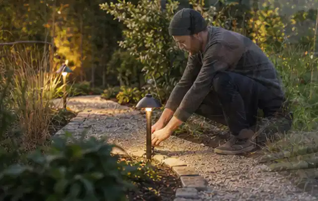 Person richtet eine niedrige warm leuchtende Gartenlampe am Kiesweg aus; Thema Mähroboter ohne Begrenzungskabel, Schwerpunkt Checkliste; Ausgangslage.