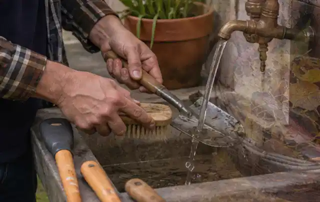 Hände reinigen gebrauchte Gartengeräte mit Bürste und Wasser am Außenbecken; Thema Mähroboter ohne Begrenzungskabel, Schwerpunkt Checkliste; Praxisalltag.