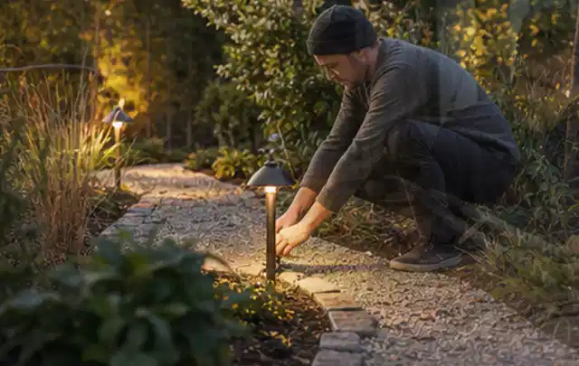 Person richtet eine niedrige warm leuchtende Gartenlampe am Kiesweg aus; Thema Mähroboter ohne Begrenzungskabel, Schwerpunkt Dimensionierung; Umsetzung.