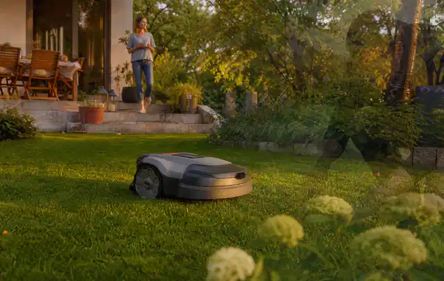 Mähroboter fährt am Abend über einen Rasen im Familiengarten; Thema Mähroboter ohne Begrenzungskabel, Schwerpunkt Ersatzteile; Ergebnis.
