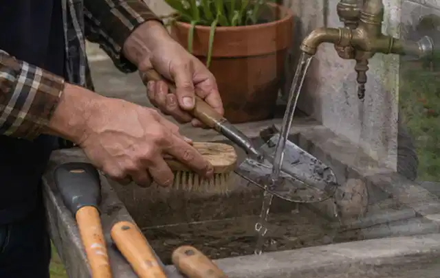 Hände reinigen gebrauchte Gartengeräte mit Bürste und Wasser am Außenbecken; Thema Mähroboter ohne Begrenzungskabel, Schwerpunkt Fehlerhilfe; Kontrolle.