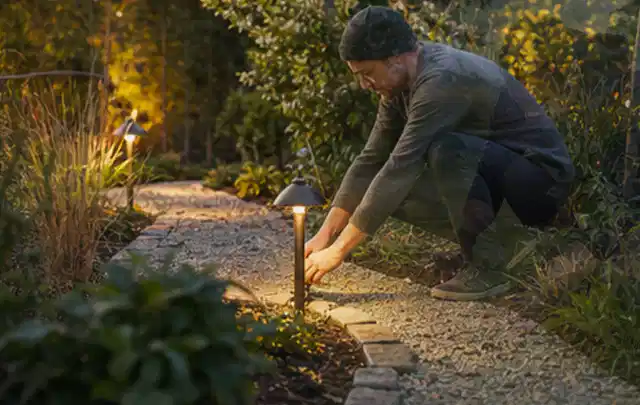 Person richtet eine niedrige warm leuchtende Gartenlampe am Kiesweg aus; Thema Mähroboter ohne Begrenzungskabel, Schwerpunkt Gärten mit kleiner Fläche;…