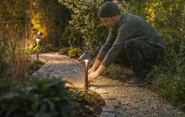 Person richtet eine niedrige warm leuchtende Gartenlampe am Kiesweg aus; Thema Mähroboter ohne Begrenzungskabel, Schwerpunkt Kaufberatung; Praxisalltag.