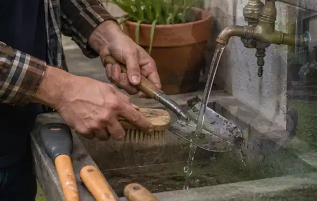 Hände reinigen gebrauchte Gartengeräte mit Bürste und Wasser am Außenbecken; Thema Mähroboter ohne Begrenzungskabel, Schwerpunkt Kosten; Ausgangslage.