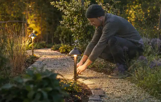 Person richtet eine niedrige warm leuchtende Gartenlampe am Kiesweg aus; Thema Mähroboter ohne Begrenzungskabel, Schwerpunkt Nachhaltigkeit; Praxisalltag.