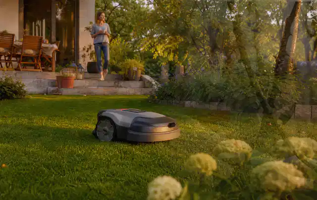 Mähroboter fährt am Abend über einen Rasen im Familiengarten; Thema Mähroboter ohne Begrenzungskabel, Schwerpunkt Sicherheit; Umsetzung.