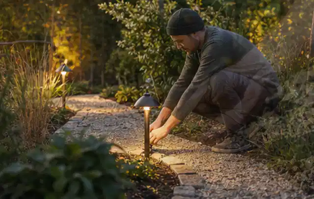Person richtet eine niedrige warm leuchtende Gartenlampe am Kiesweg aus; Thema Mähroboter ohne Begrenzungskabel, Schwerpunkt Systemvergleich; Umsetzung.