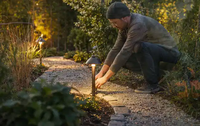 Person richtet eine niedrige warm leuchtende Gartenlampe am Kiesweg aus; Thema Mähroboter ohne Begrenzungskabel, Schwerpunkt Testkriterien; Kontrolle.