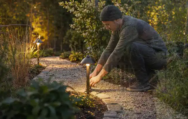 Person richtet eine niedrige warm leuchtende Gartenlampe am Kiesweg aus; Thema Mähroboter ohne Begrenzungskabel, Schwerpunkt Vergleich; Kontrolle.