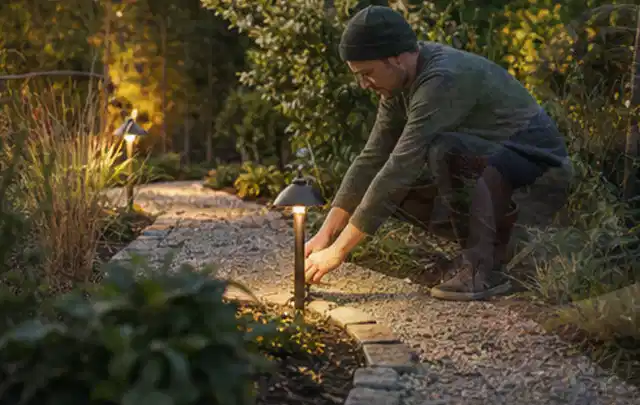 Person richtet eine niedrige warm leuchtende Gartenlampe am Kiesweg aus; Thema Mähroboter ohne Begrenzungskabel, Schwerpunkt Wartung; Umsetzung.