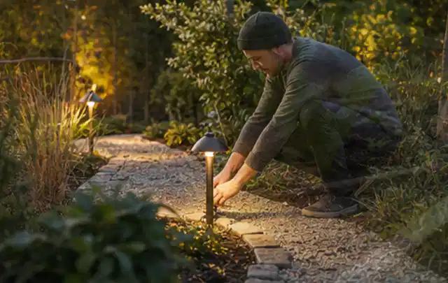 Person richtet eine niedrige warm leuchtende Gartenlampe am Kiesweg aus; Thema Mammotion Luba, Schwerpunkt Dimensionierung; Kontrolle.
