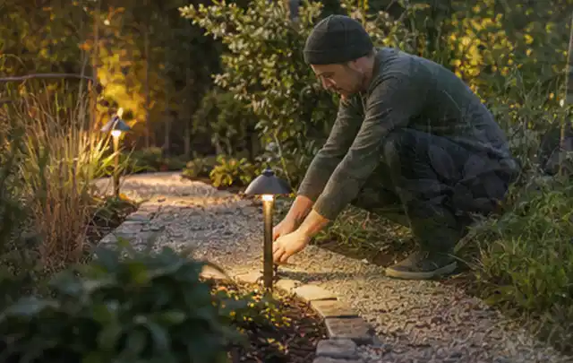 Person richtet eine niedrige warm leuchtende Gartenlampe am Kiesweg aus; Thema Mammotion Luba, Schwerpunkt Gärten mit großer Fläche; Praxisalltag.