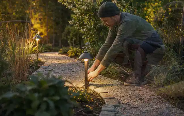 Person richtet eine niedrige warm leuchtende Gartenlampe am Kiesweg aus; Thema Mammotion Luba, Schwerpunkt Nachhaltigkeit; Kontrolle.