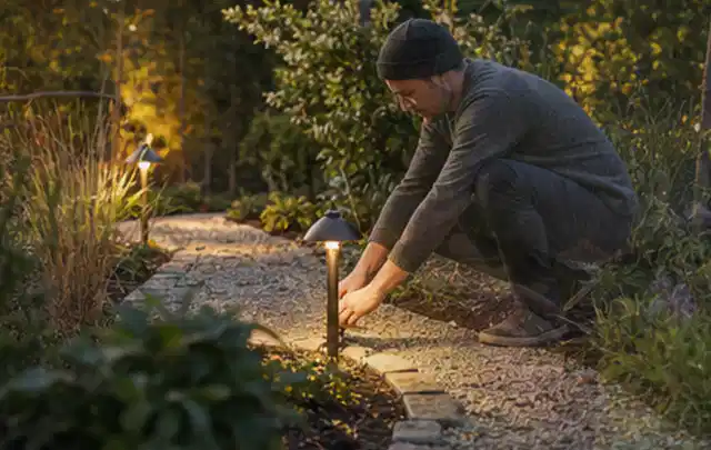 Person richtet eine niedrige warm leuchtende Gartenlampe am Kiesweg aus; Thema Mammotion Luba, Schwerpunkt Sicherheit; Praxisalltag.