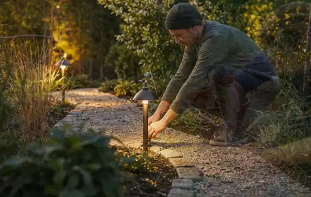 Person richtet eine niedrige warm leuchtende Gartenlampe am Kiesweg aus; Thema Mammotion Luba, Schwerpunkt Systemvergleich; Praxisalltag.