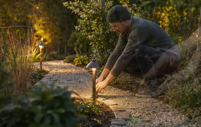 Person richtet eine niedrige warm leuchtende Gartenlampe am Kiesweg aus; Thema Mammotion Luba, Schwerpunkt Wartung; Kontrolle.