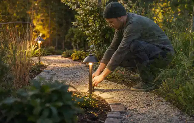 Person richtet eine niedrige warm leuchtende Gartenlampe am Kiesweg aus; Thema Marken, Schwerpunkt Mähroboter; Kontrolle.