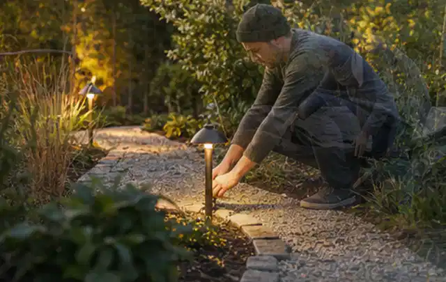 Person richtet eine niedrige warm leuchtende Gartenlampe am Kiesweg aus; Thema Mediterraner Garten, Schwerpunkt Bepflanzung; Umsetzung.